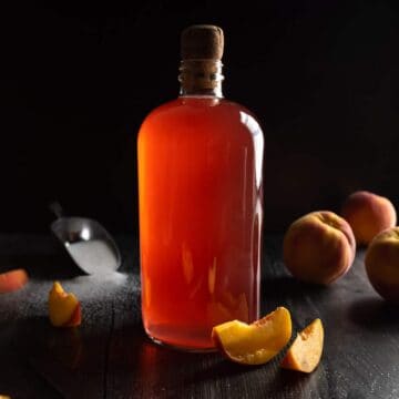 A corked glass bottle of peach syrup surrounded by whole and sliced peaches.