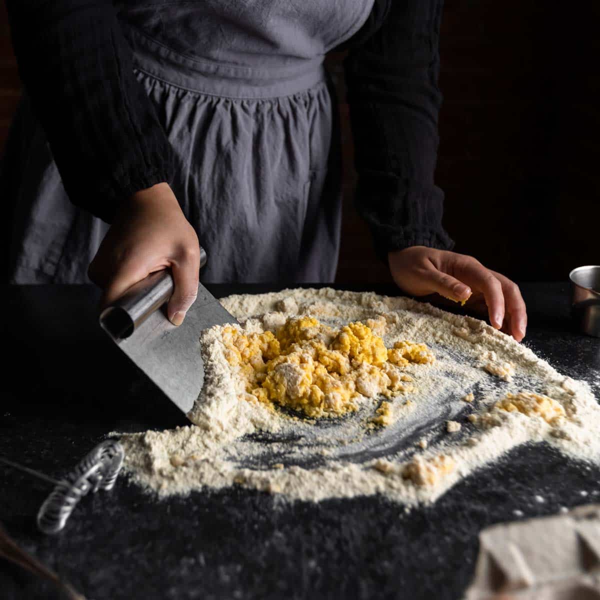 A woman using a bench scraper to incorporate flour into fresh pasta dough.