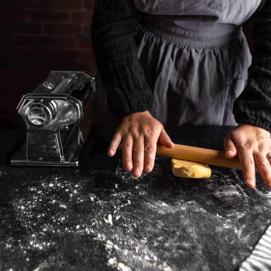 A woman rolling a piece of pasta dough with a small rolling pin on a floured surface