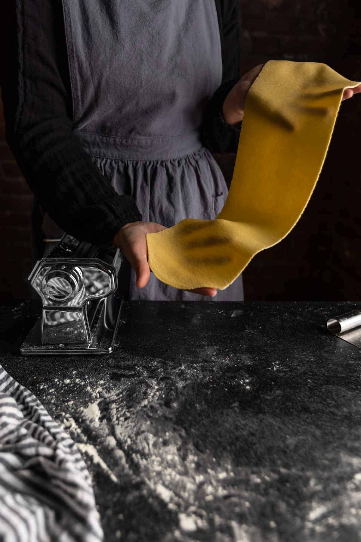 An aproned woman holding up a thin translucent sheet of semolina pasta dough 