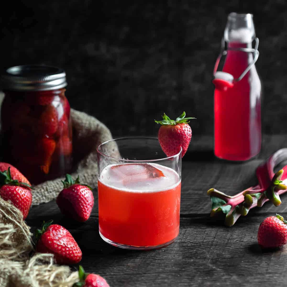 A strawberry rhubarb whiskey sour with a strawberry garnish. A jar of strawberry infused whiskey, fresh strawberries, stalks of rhubarb and a bottle of rhubarb syrup are in the background.