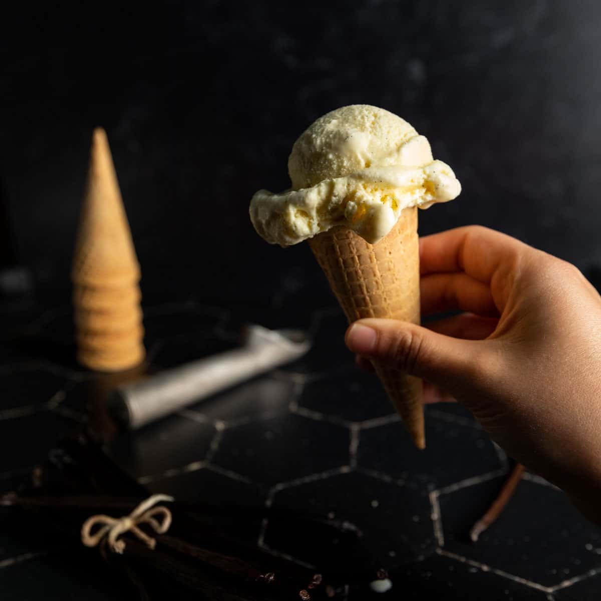 A hand holding a sugar cone topped with one scoop of vanilla ice cream