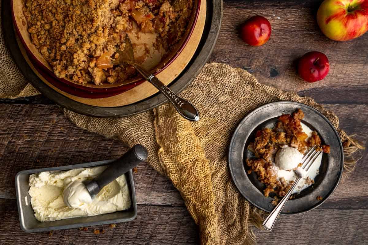 A scoop of vanilla bean ice cream melting on top of a serving of plum and apple crumble next to the baking dish, ice cream and ice cream scoop