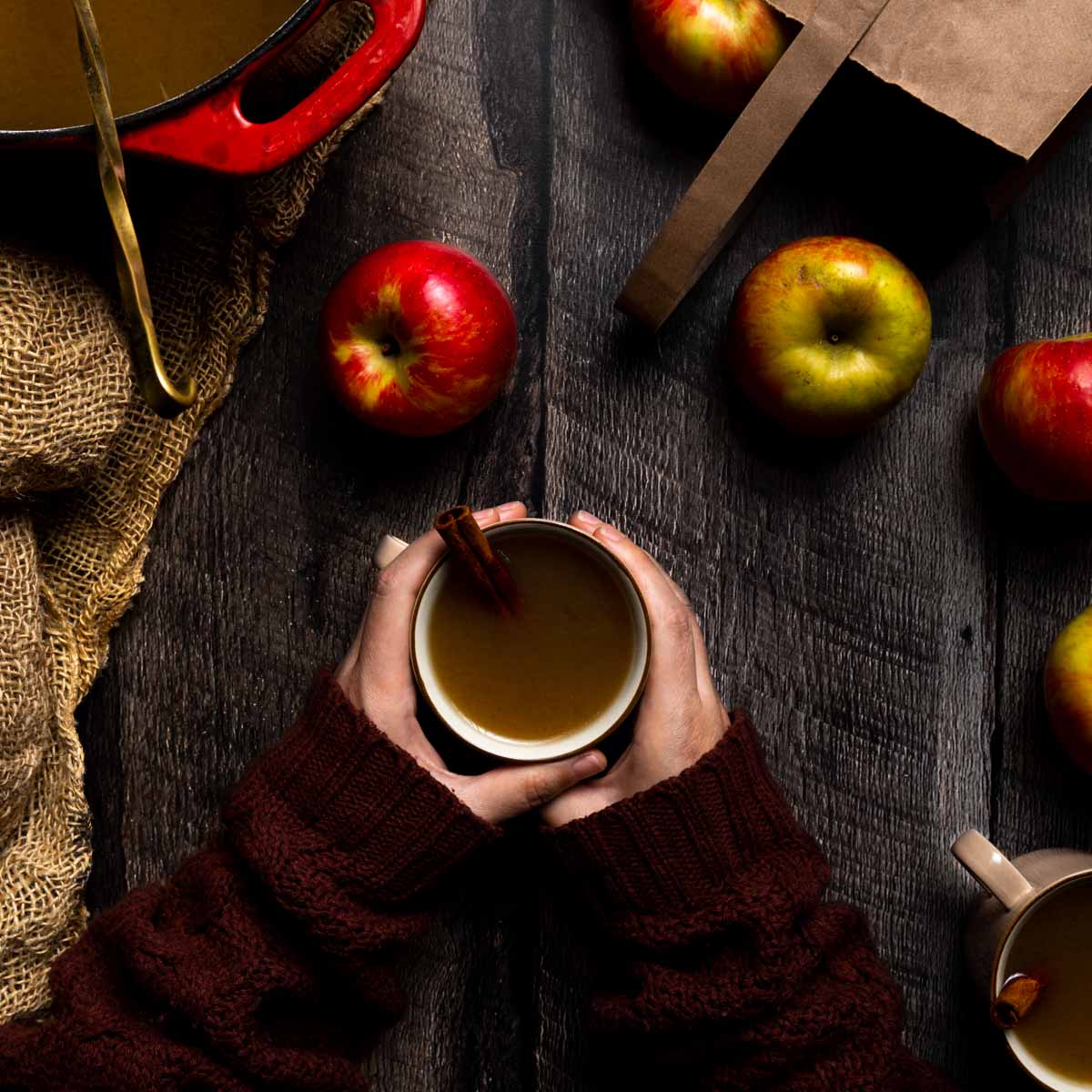 Two hands cradling a mug of homemade spiced cider next to apples and burlap