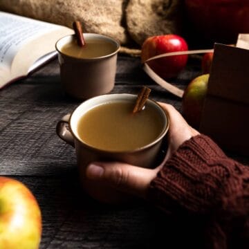 A hand holding a mug of hot apple cider next to apples, books, and another mug