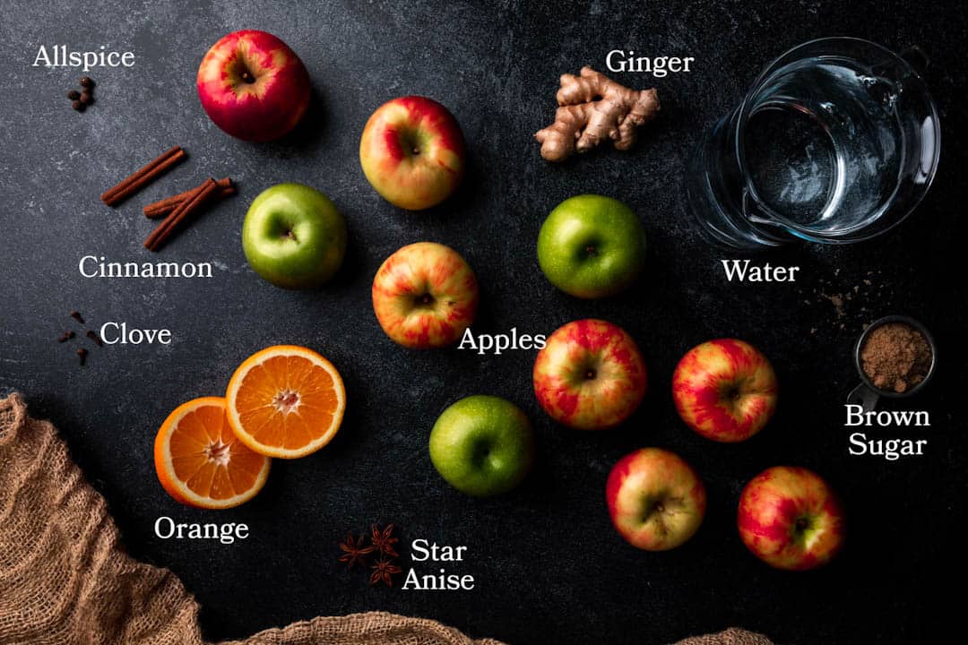 Picture of ingredients for homemade apple cider including apples, orange, ginger, brown sugar, star anise, clove, cinnamon, allspice and water. 