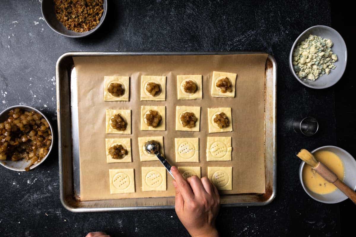 A hand putting heaping teaspoons of ear chutney on each piece of puff pastry on a parchment lined baking sheet.