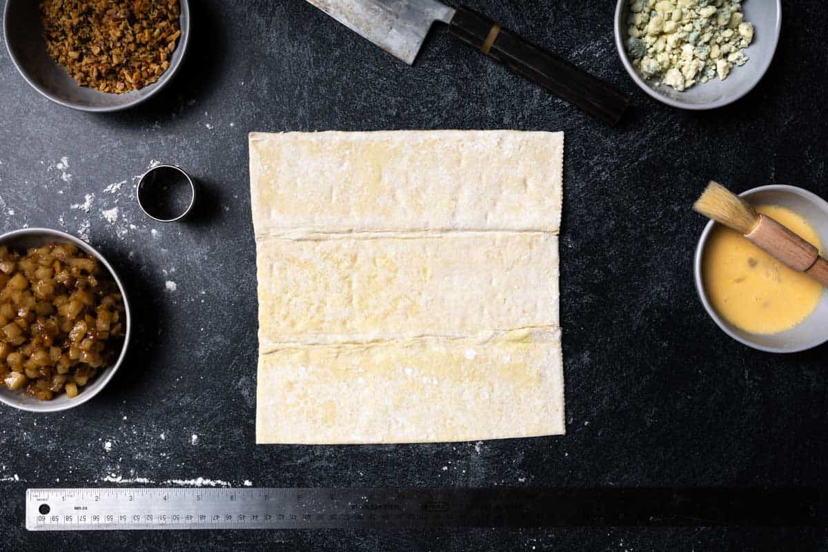 A piece of puff pastry dough with a ruler and bowls of ingredients