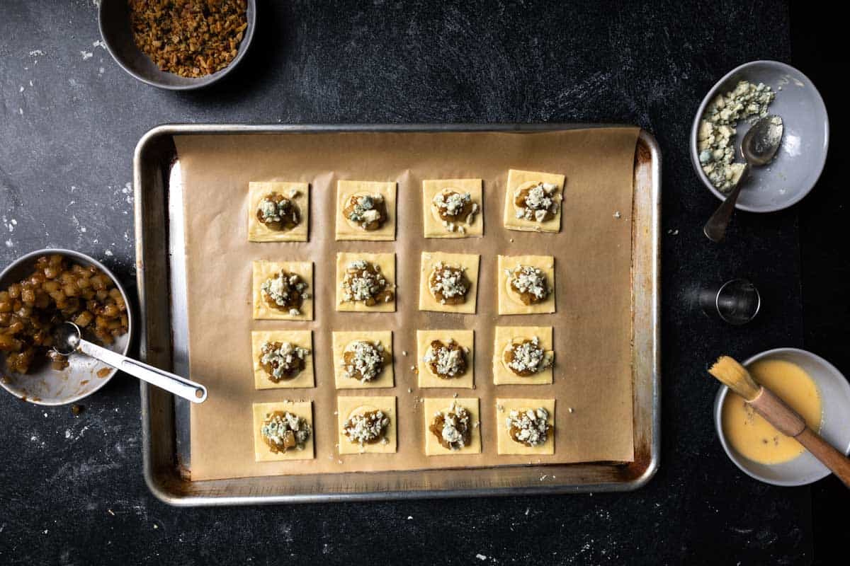 A hand using a small spoon to top each pastry square with a sprinkle of gorgonzola cheese