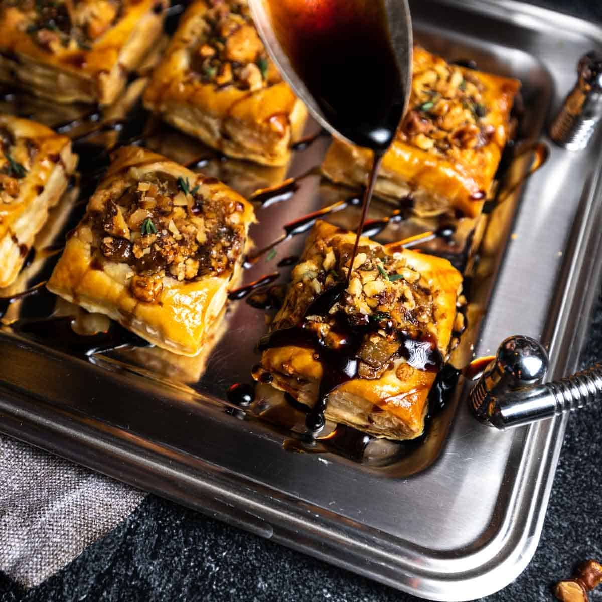 A spoon drizzling balsamic glaze over a pear and blue cheese appetizer tart.
