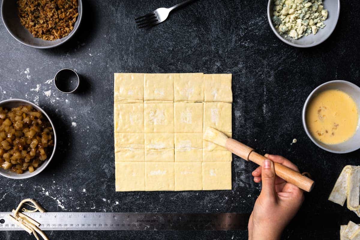 A hand holding a pastry brush and brushing the cut edges of the puff pastry dough with egg wash