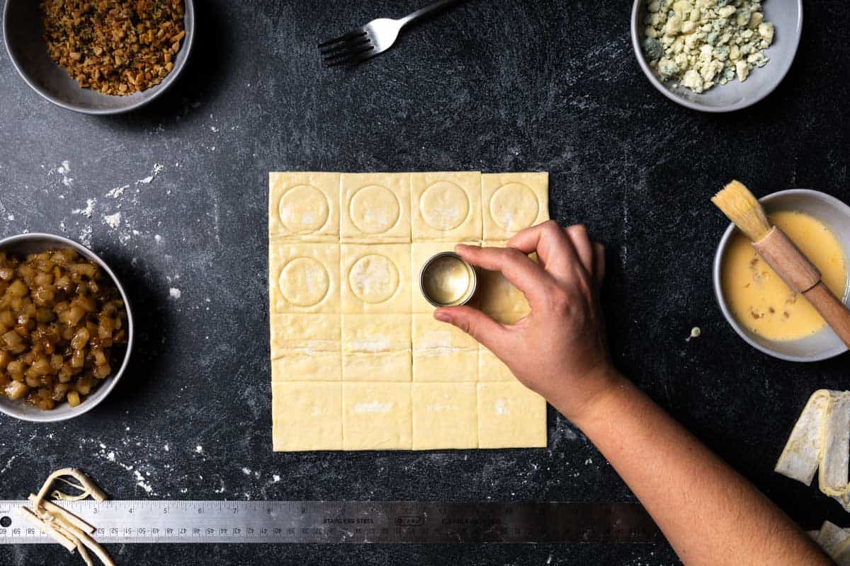 A hand using a small circular cookie cutter to score the center of each puff pastry square