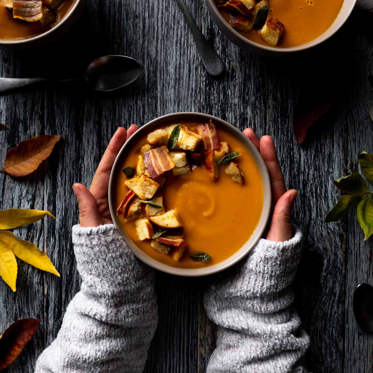 Arms in a knitted sweater cradling a big bowl of sweet potato and pumpkin soup topped with croutons, sage leaves and pancetta.