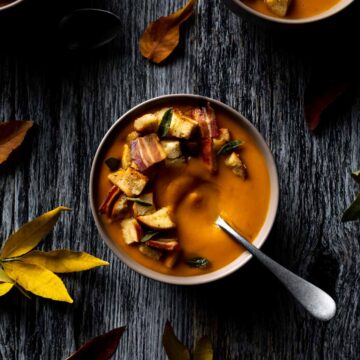 A spoon in a big bowl of pumpkin sweet potato soup topped with pancetta, sage and croutons