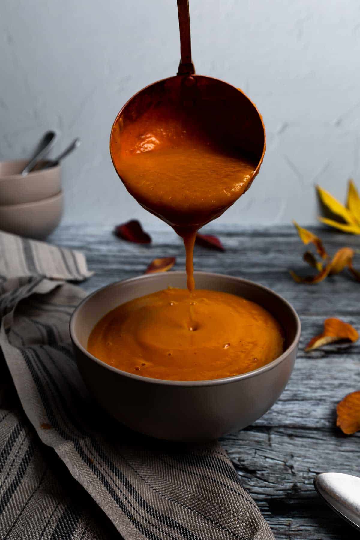 A big copper ladle pouring roasted pumpkin and sweet potato soup into a bowl surrounded by colorful autumn leaves