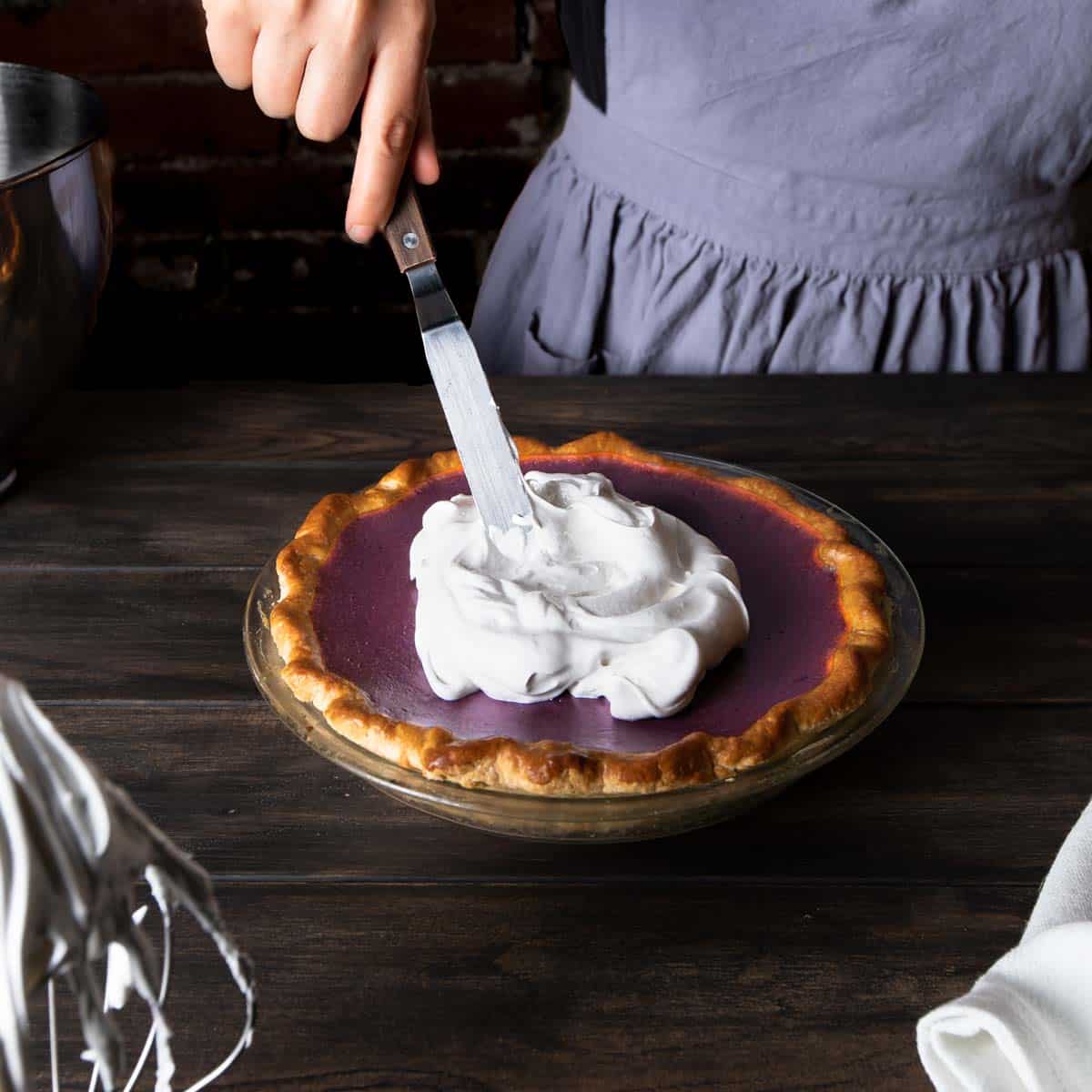 A woman in an apron spreading Swiss meringue on top of a baked sweet potato pie with an offset spatula