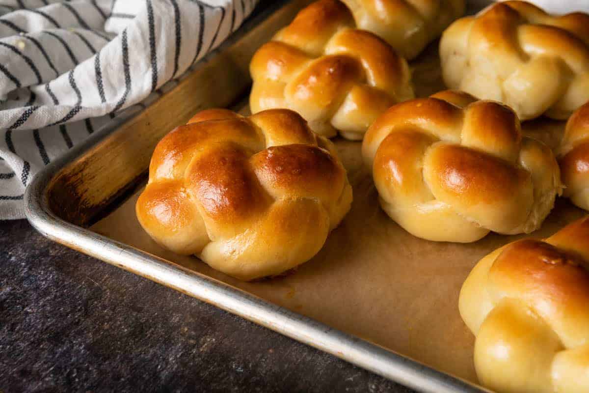 Several challah bread rolls lined up on a baking tray next to a striped linen.