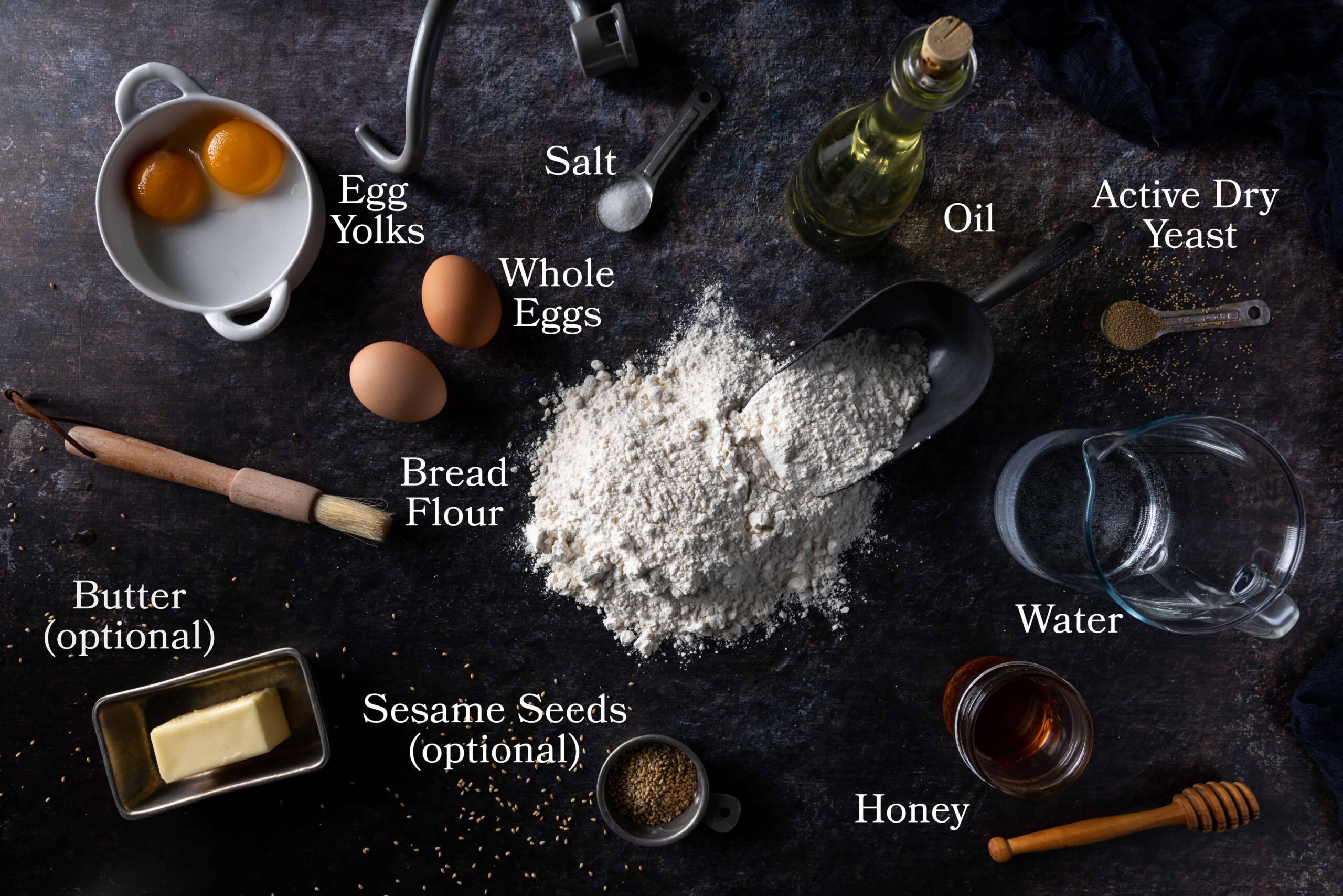 Ingredients for challah buns including bread flour, salt, yeast, honey, oil, eggs, egg yolks, water and optional sesame seeds and butter