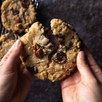 A hand breaking a big gooey dark chocolate coconut pecan cookie in half