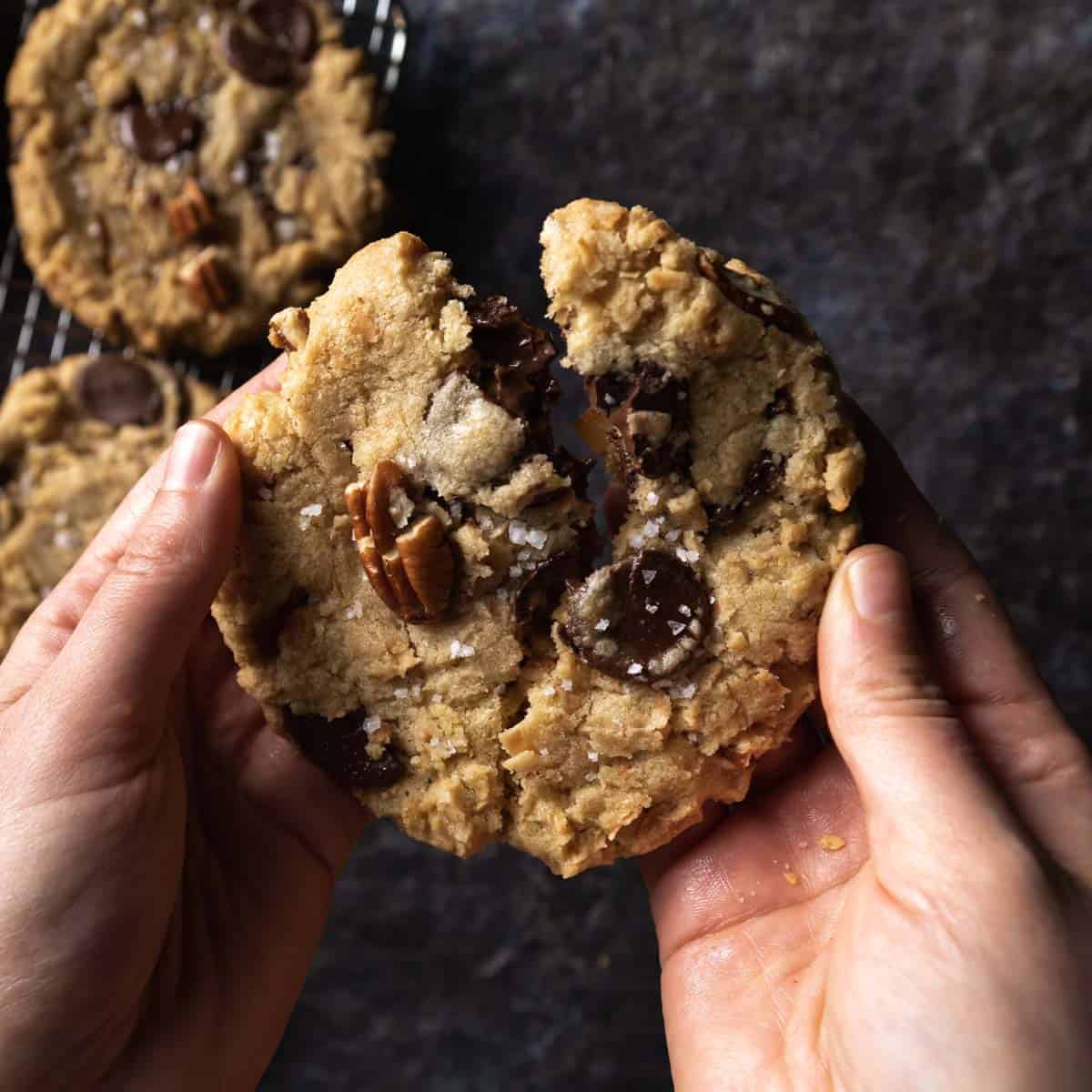 A hand breaking a big gooey dark chocolate coconut pecan cookie in half