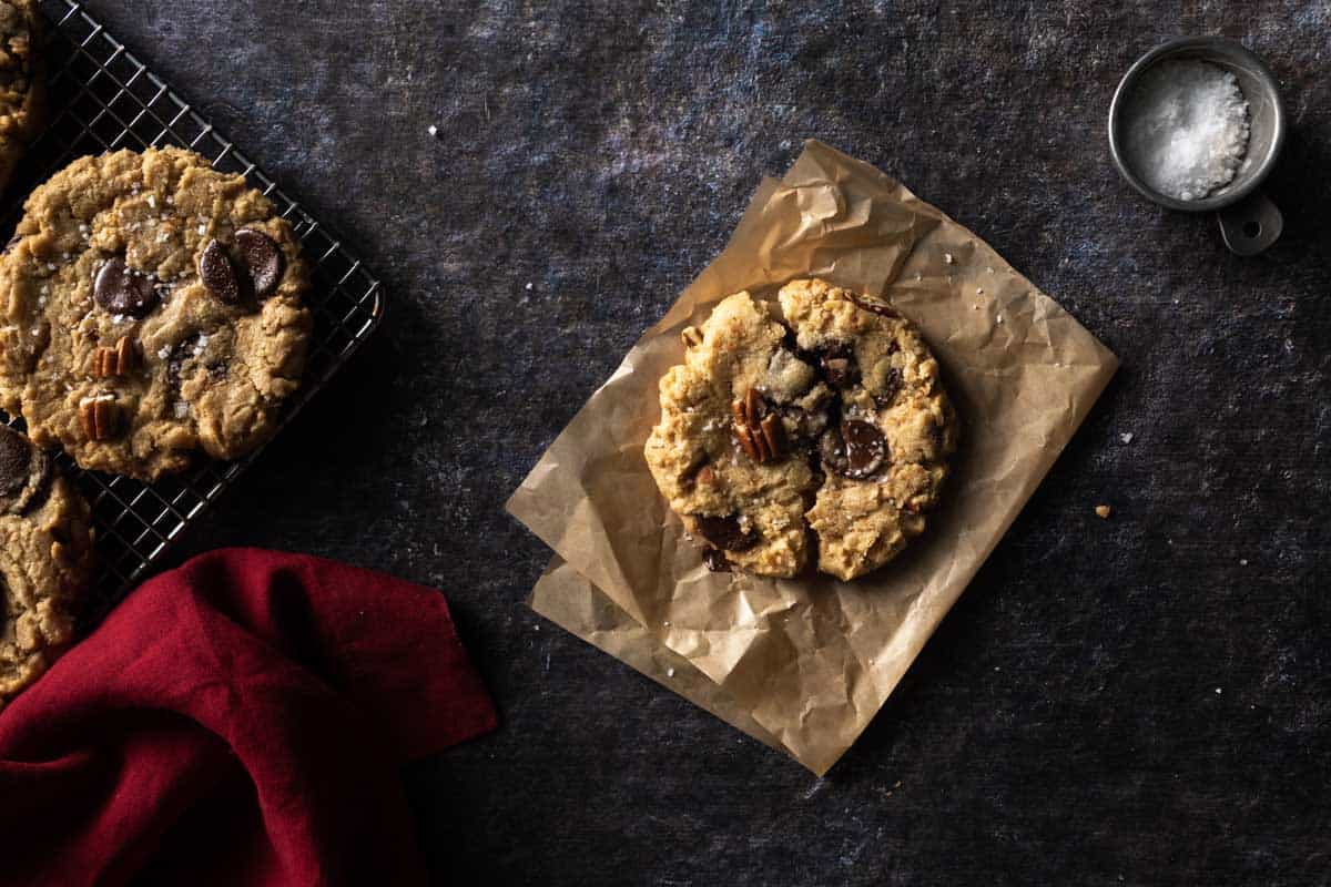 A broken coconut pecan chocolate chunk cookie on a piece of crumpled parchment paper next to a cooling rack full of cookies