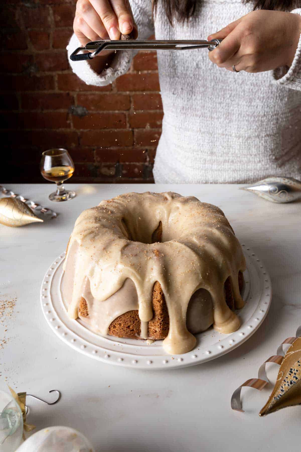 A woman in a white sweater grating fresh nutmeg over an eggnog cake with rum icing