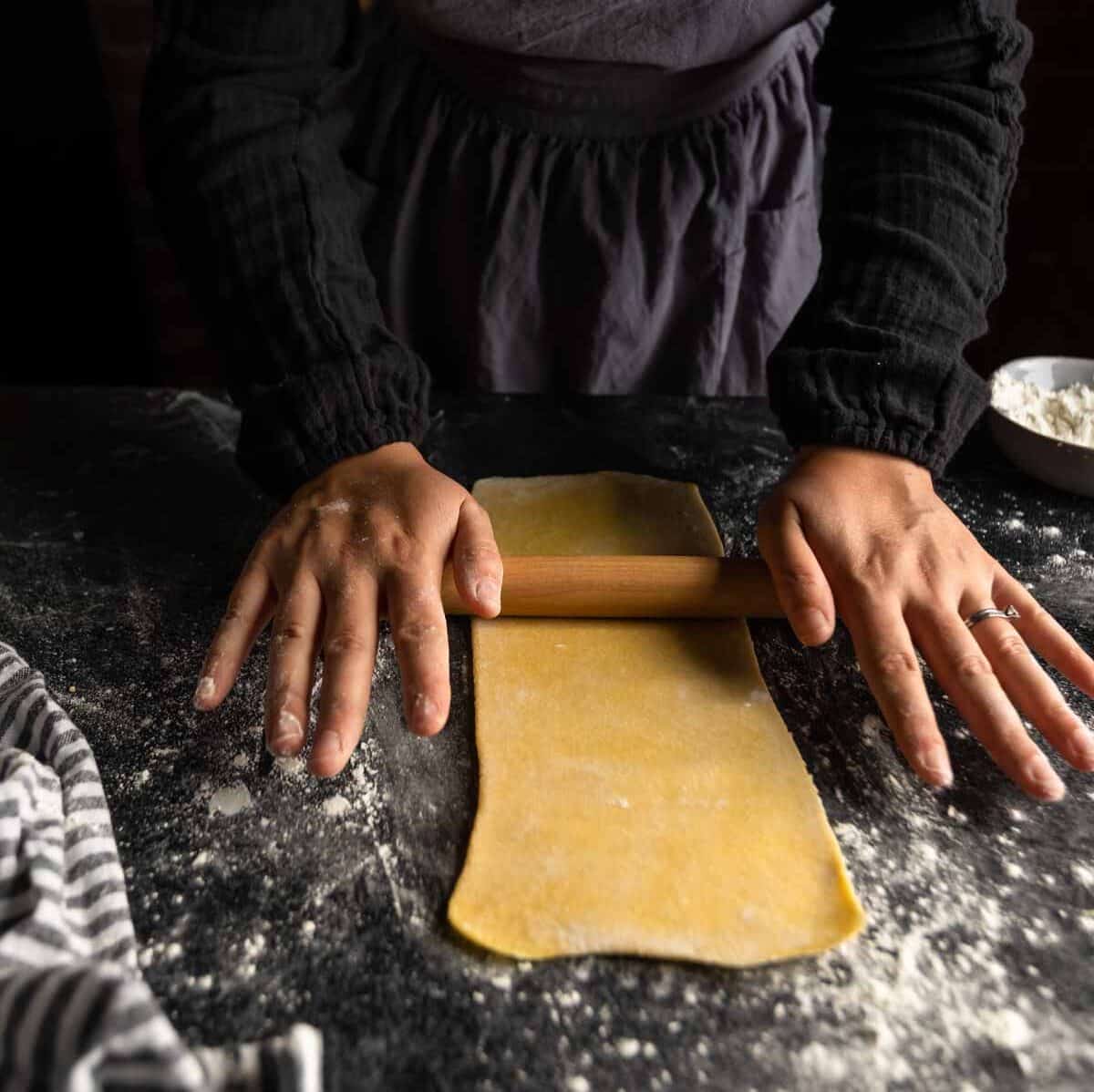 A woman rolling a long thin rectangle of pasta dough with a small rolling pin.