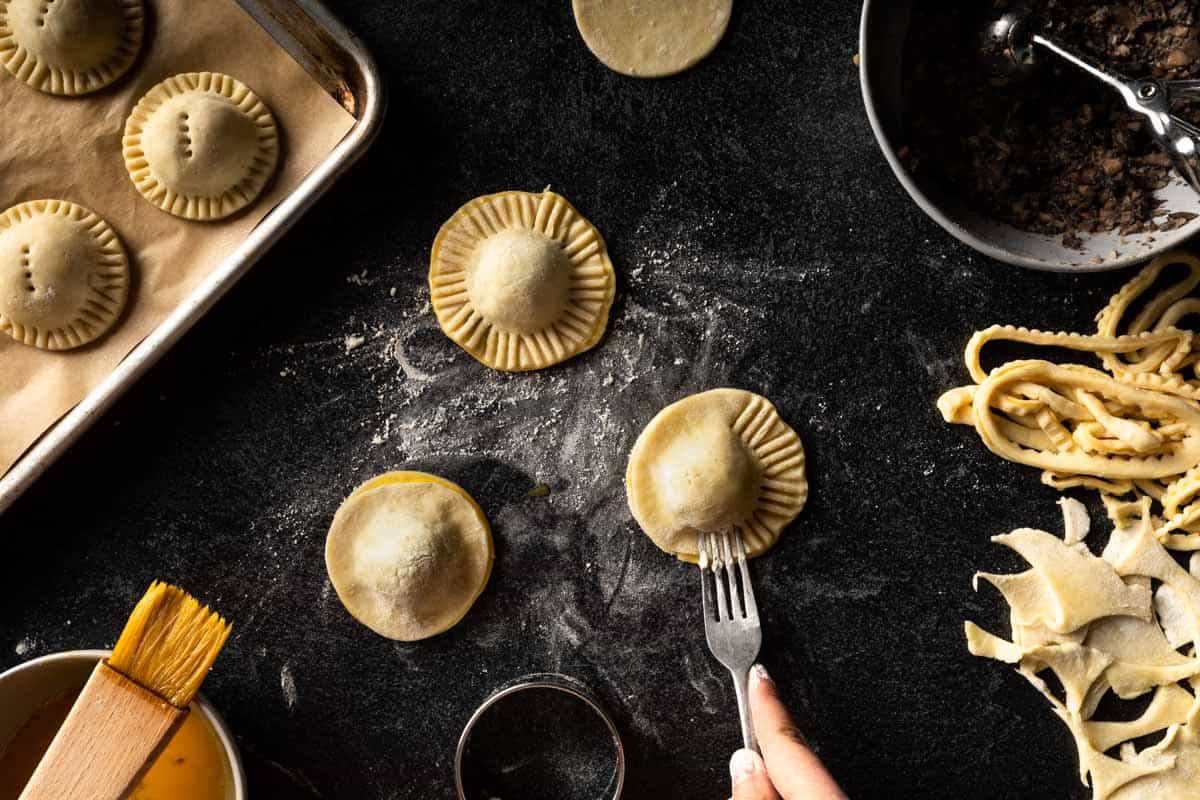 A busy scene of mini pie making including a fork crimping the edges of a mini mushroom pie.