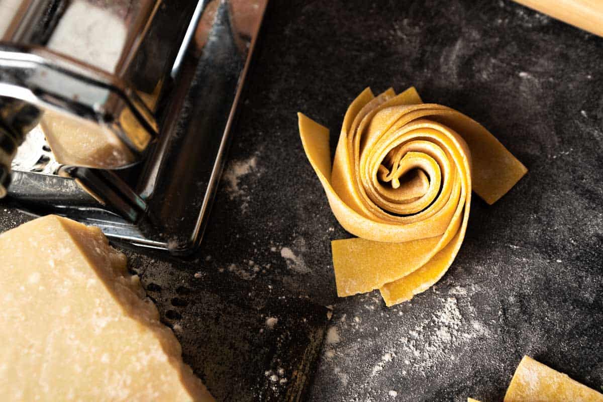 A spiraled nest of fresh pappardelle on a floured surface next to a pasta machine and wedge of Parmesan cheese