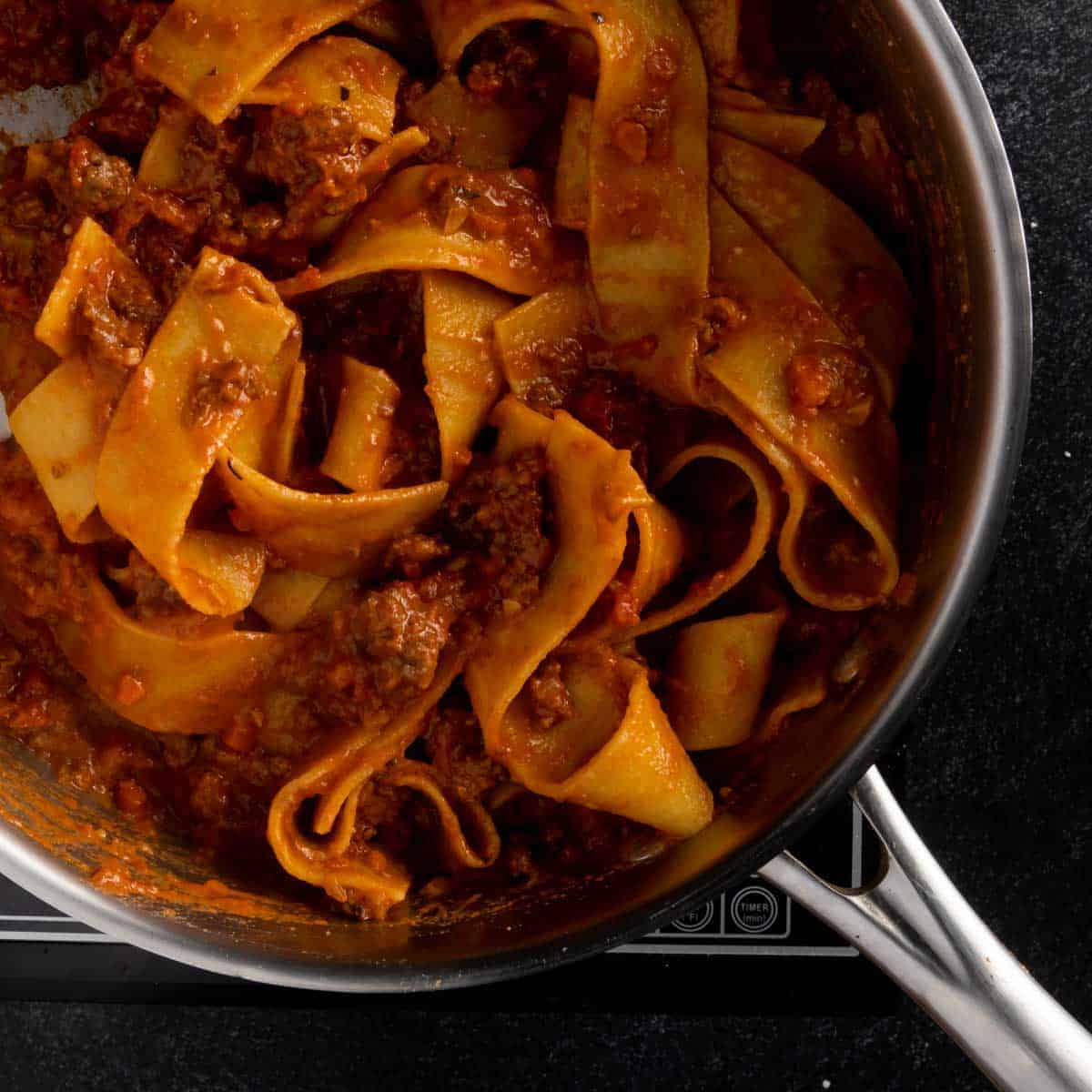 Fresh pappardelle pasta being cooked in a skillet with ragu alla Bolognese and pasta water