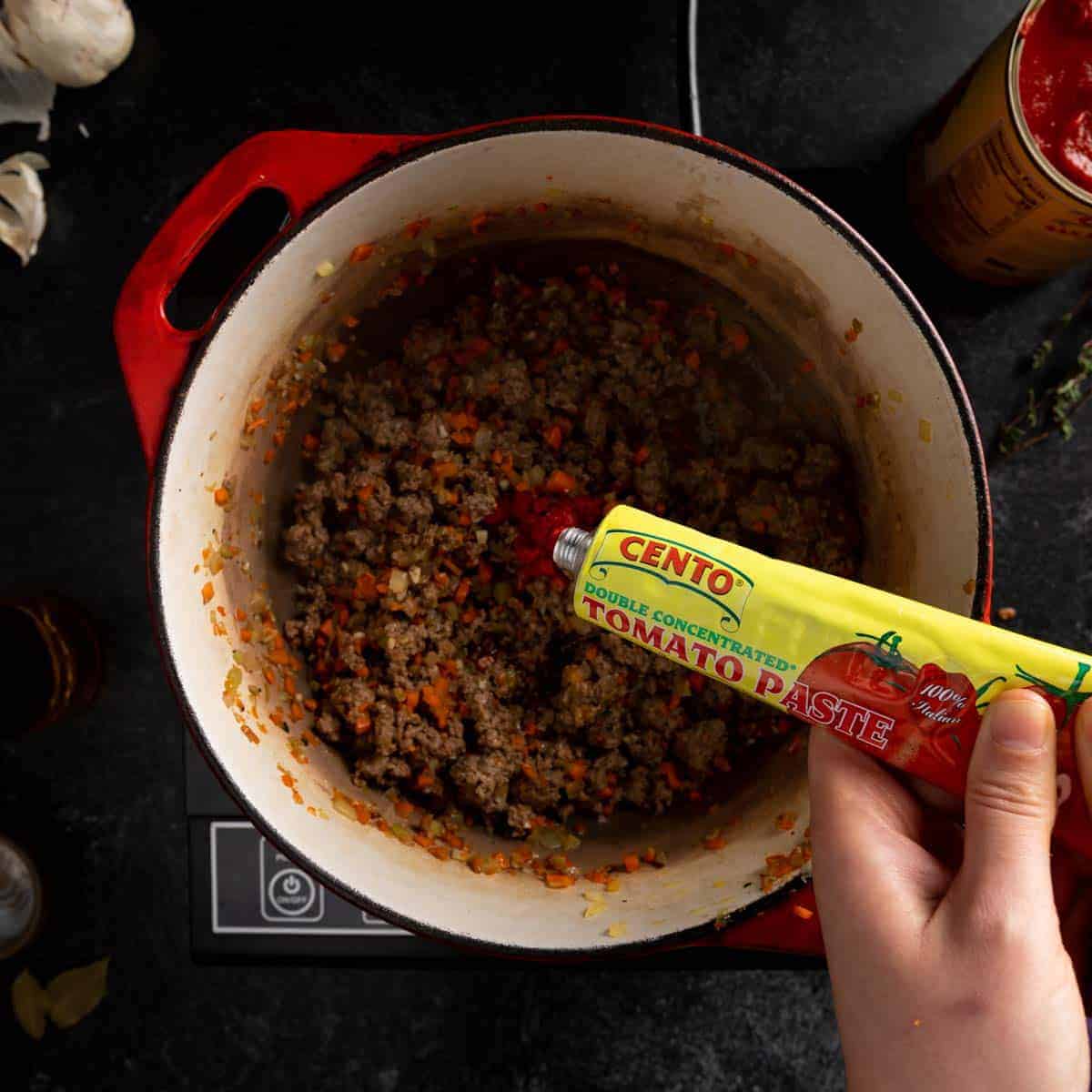 Squeezing a tube of tomato paste into the dutch oven with the cooked ground beef and soffrito.