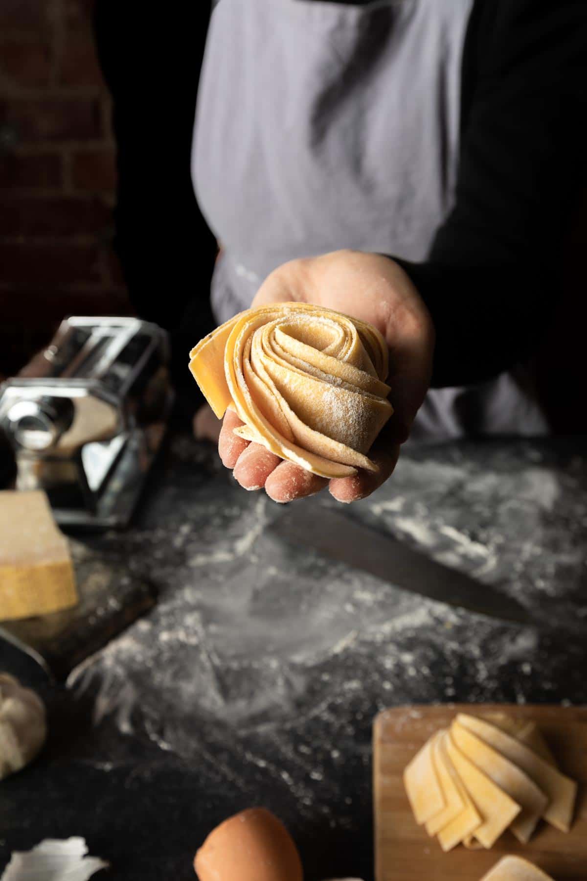 A woman holding out a nest of Italian pappardelle pasta over a floured work surface. 