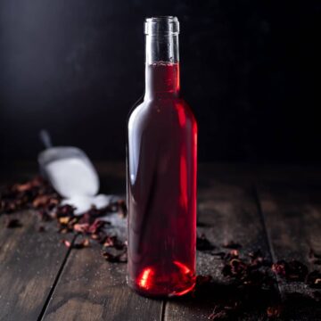 A glass jar full of hibiscus syrup surrounded by a scoop of sugar and dried hibiscus flowers