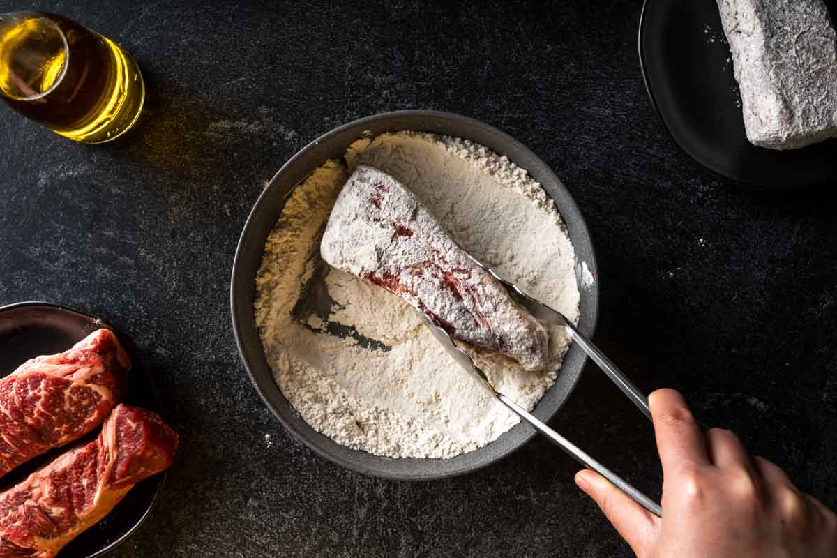 Dredging a raw boneless beef short rib through seasoned flour with tongs