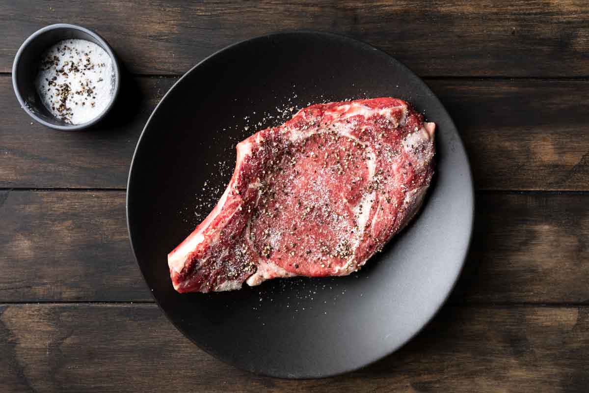 A raw ribeye steak seasoned with kosher salt and black pepper