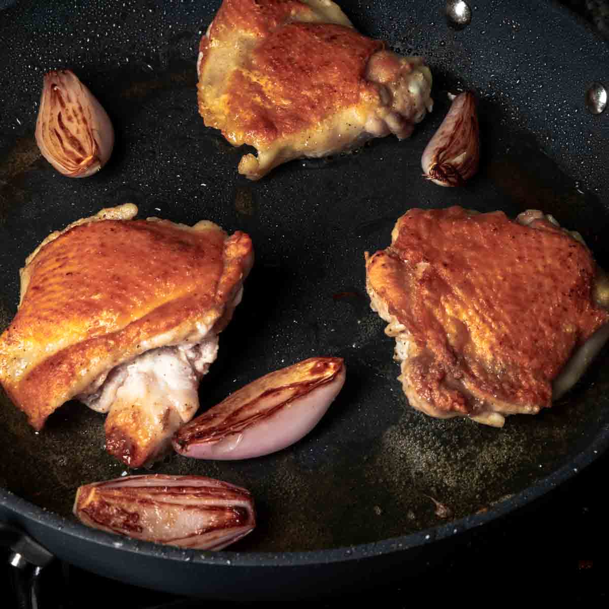 Golden brown chicken thighs and shallots in a frying pan.
