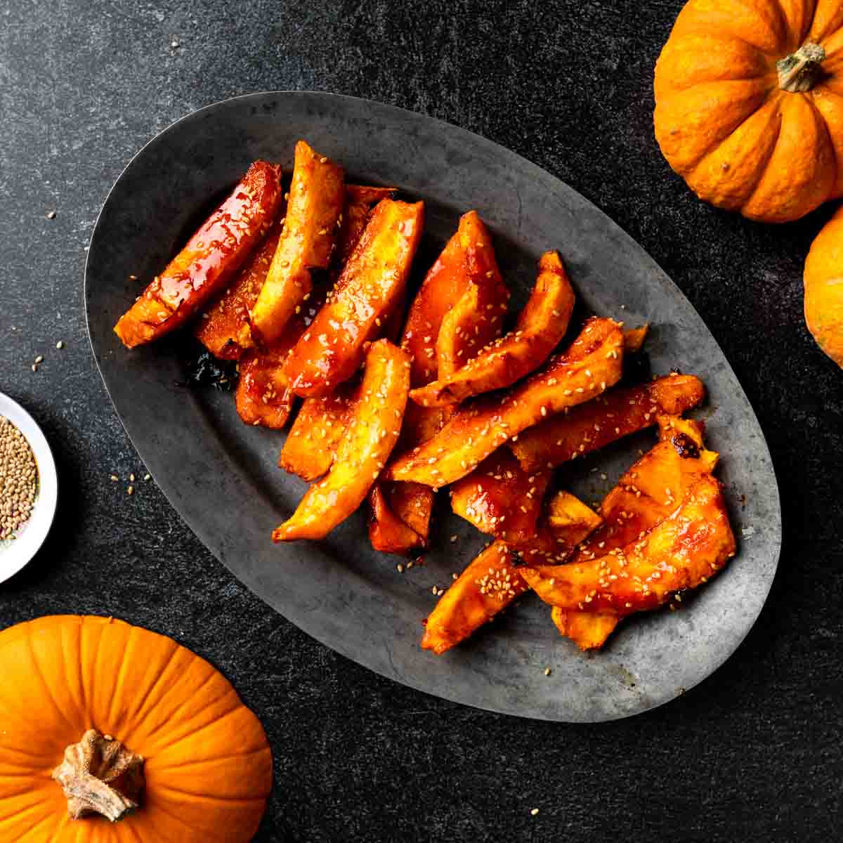 An oval dish filled with spicy maple pumpkin wedges garnished with sesame seeds.