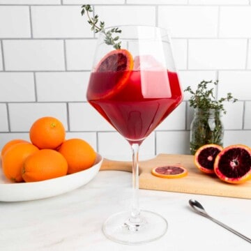 A wine glass filled with a blood orange cocktail garnished with a sprig of thyme.