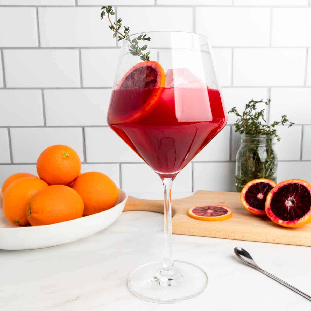 A wine glass filled with a blood orange cocktail garnished with a sprig of thyme. 