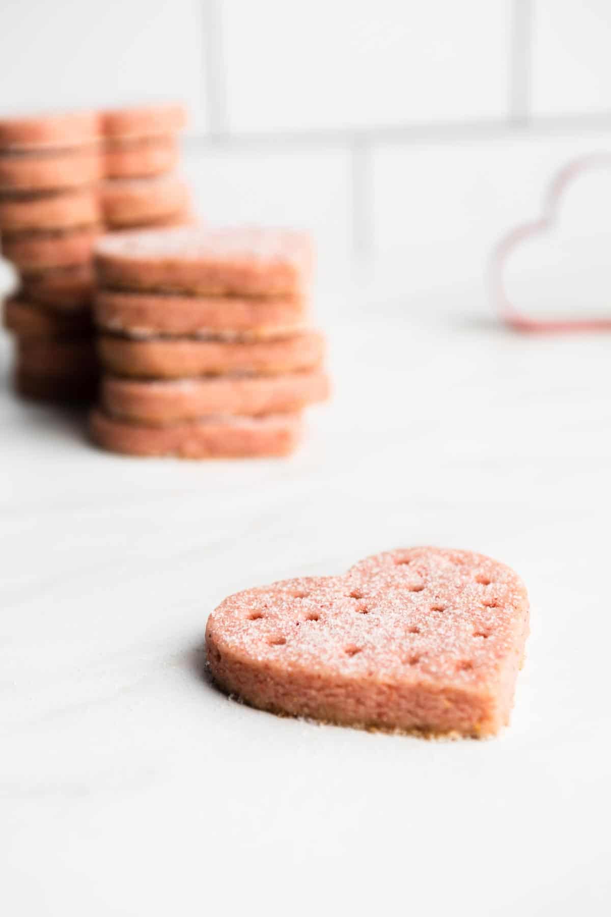 A heart shaped strawberry shortbread cookie next to stack of more cookies.