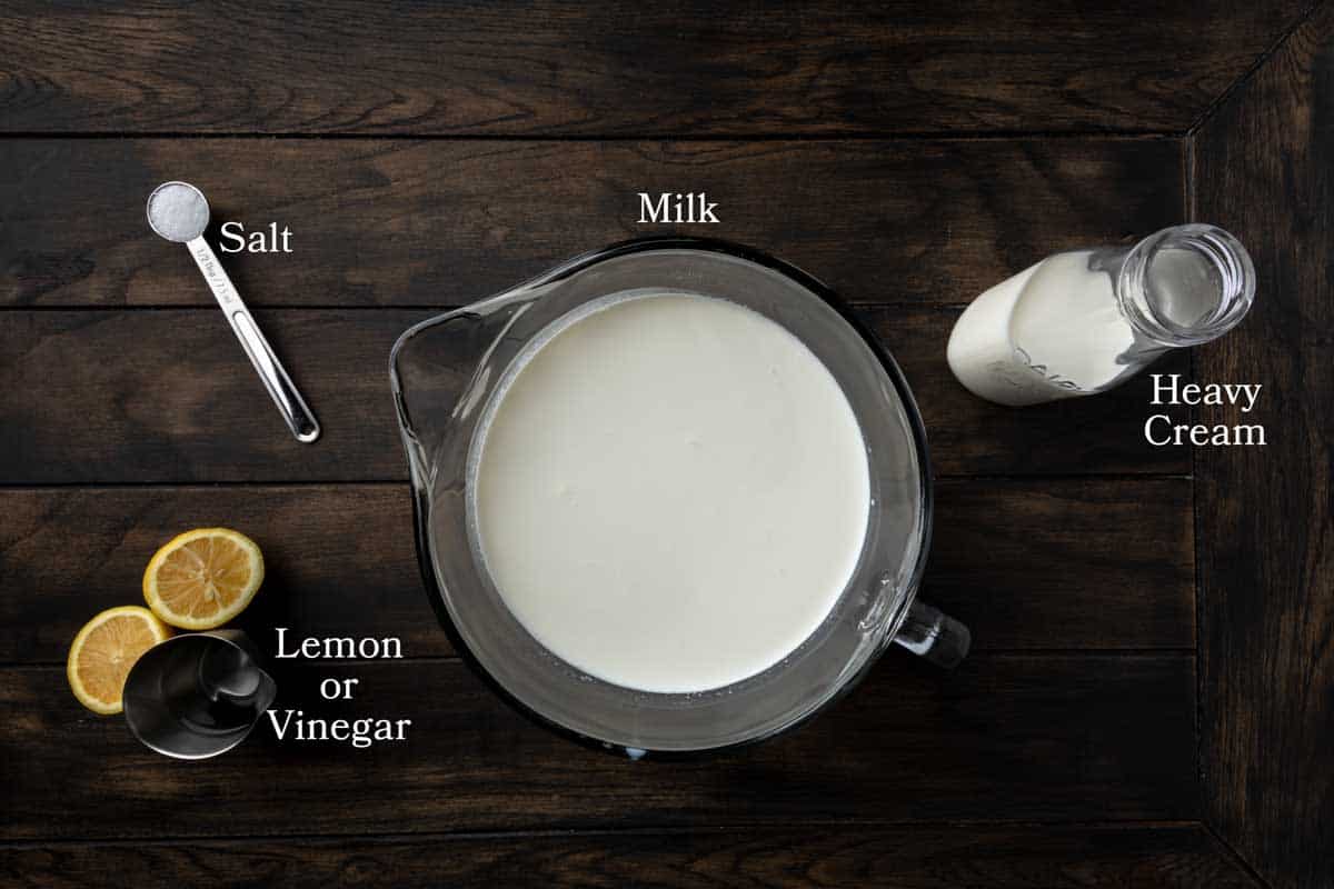 A large bowl of milk, a glass jar of heavy cream, a spoon of salt and white vinegar next to a lemon. 