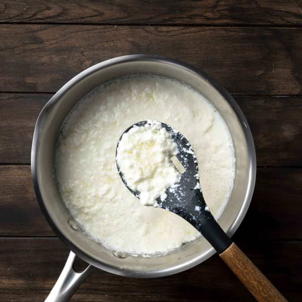 A spoon holding up separated curds of ricotta cheese over a pot of acidified milk. 