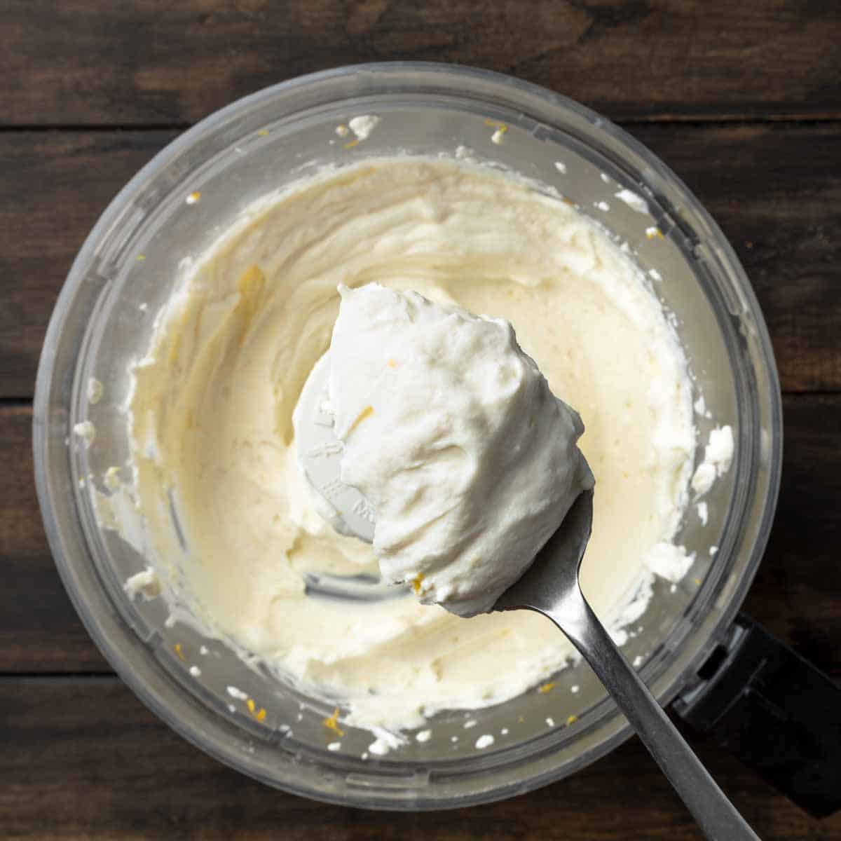 A spoonful of creamy whipped ricotta cheese with lemon zest over a food processor. 