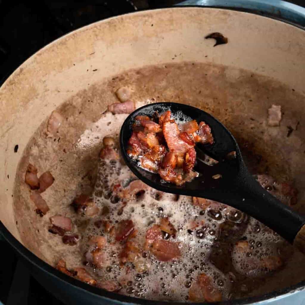 A slotted spoon removing lardons from a Dutch oven with bacon and grease.