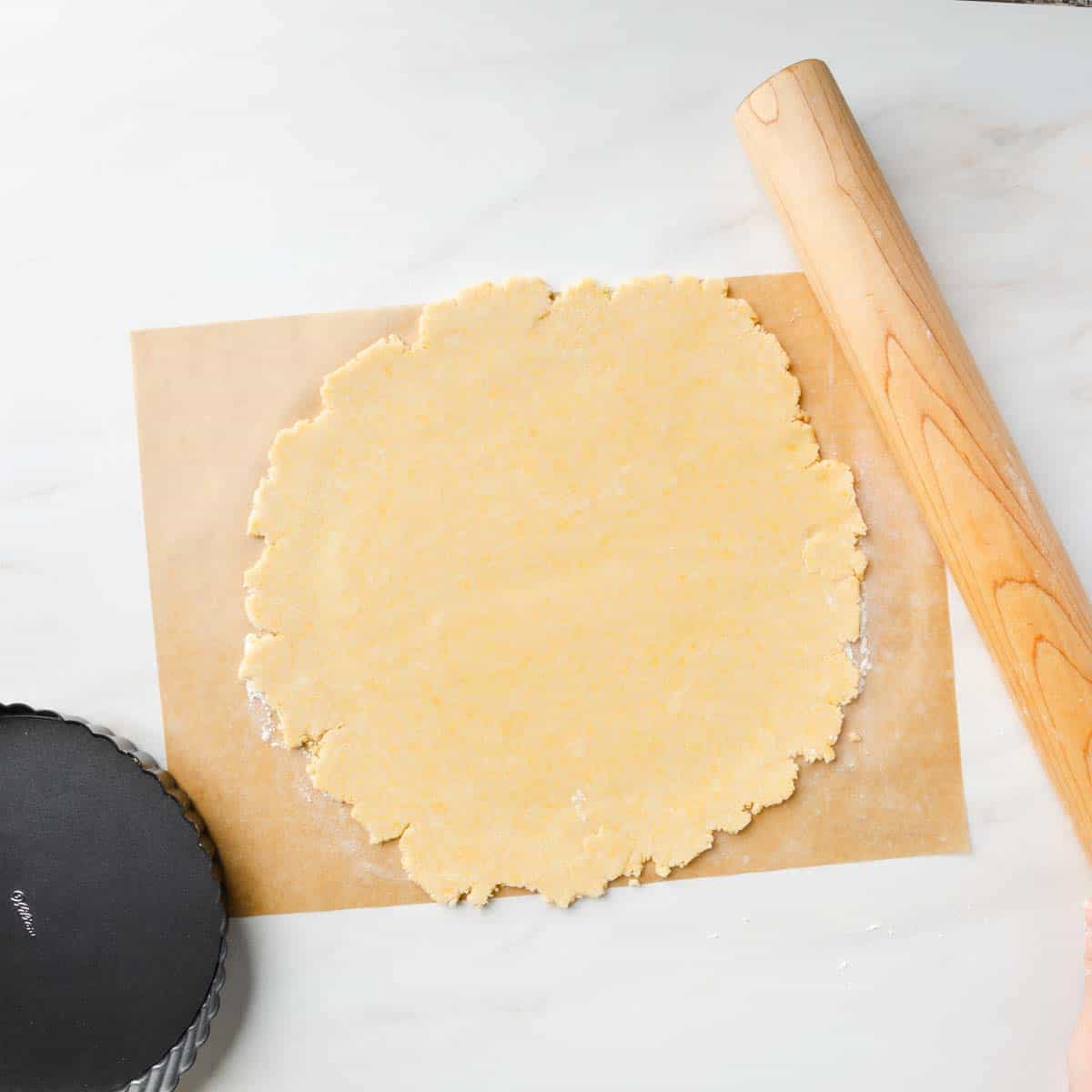 A rolled out disk of pasta frolla dough on a sheet of parchment paper next to a rolling pin.