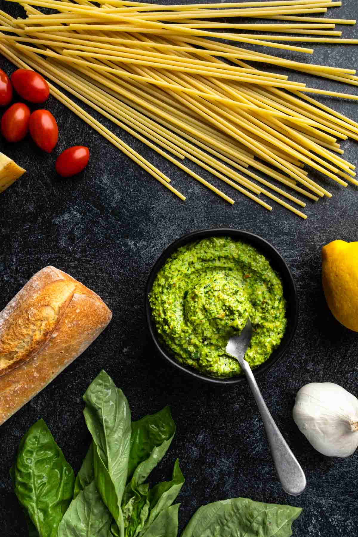 A bowl of pesto next to raw spaghetti, cherry tomatoes, garlic and a baguette. 