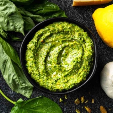 A bowl of bright green spinach pistachio pesto surrounded by lemon, garlic and basil.