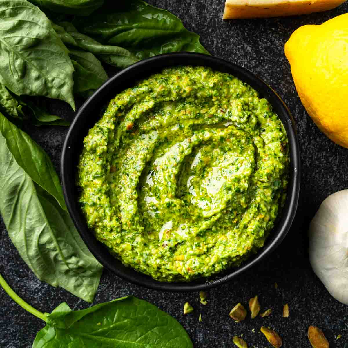 A bowl of bright green spinach pistachio pesto surrounded by lemon, garlic and basil. 