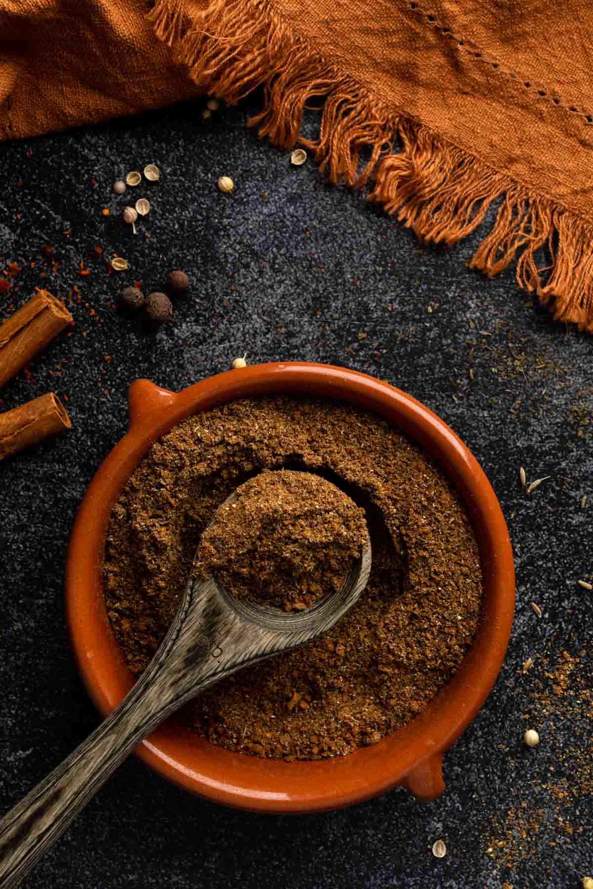 A spoonful of baharat seasoning in a terra cotta dish with a sprinkle of whole spices.