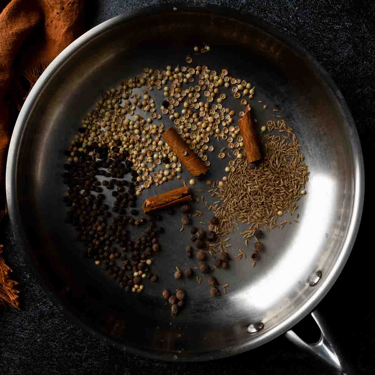 A pan full of whole toasted spices including pepper, cumin, coriander and cinnamon sticks. 